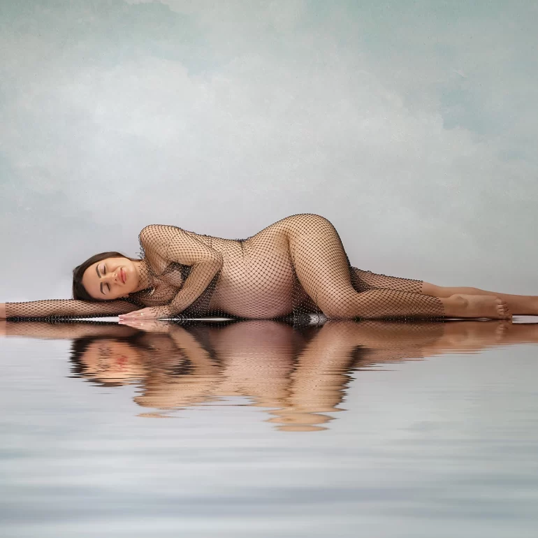 Reclining on reflective surface; shimmering mesh gown, pastel background.