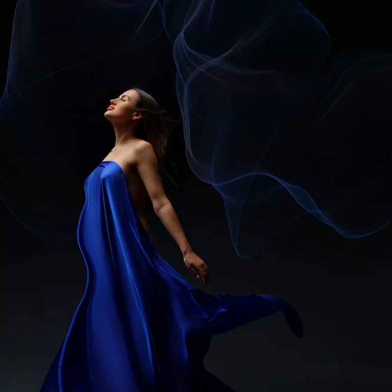 Draped in blue satin; eyes closed as wisps of light swirl around.