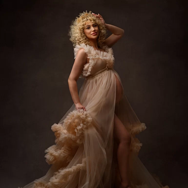 Ruffled sheer beige gown and golden crown; confident expectant mother, dramatic light.