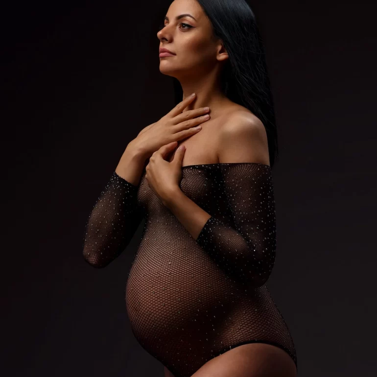 Sheer sparkling black bodysuit; side‑lit expectant mother with calm expression.
