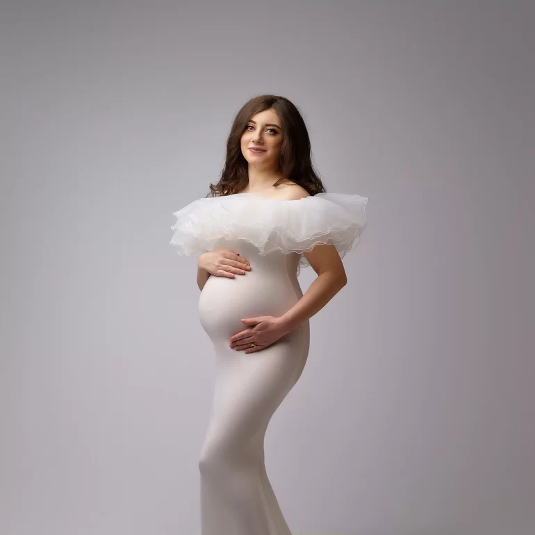 Light‑gray backdrop; off‑shoulder white ruffled gown; gentle, proud pose.