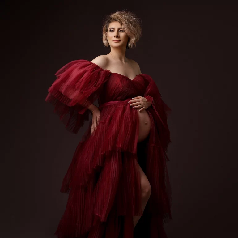 Layered burgundy gown; gentle hand on belly; dramatic studio light.