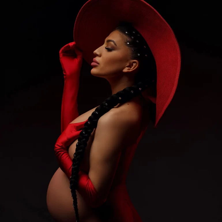 Red hat and gloves, pearl braid; bold side‑profile of pregnant woman.