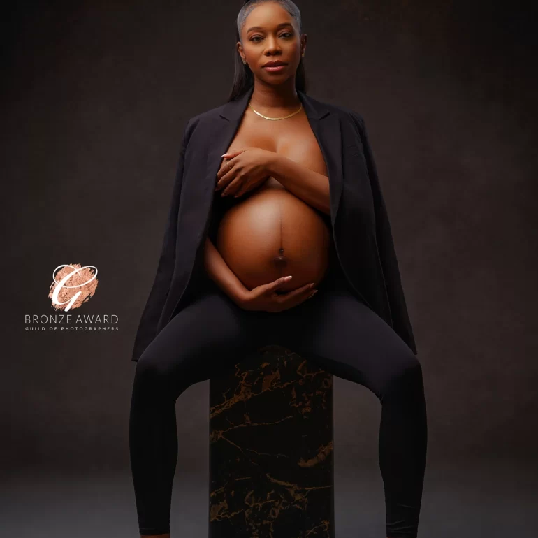 Expectant mother seated on marble pedestal in sleek black; sculpted, moody portrait.