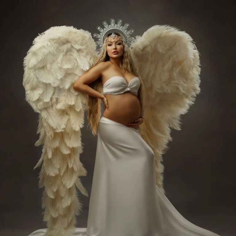 Expectant mother in white bandeau and skirt, feathered wings, crown; moody studio portrait.