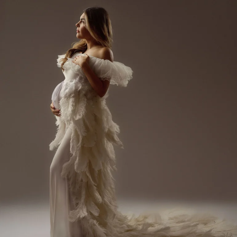 Profile in off‑shoulder white feathered gown; soft, ethereal studio scene.