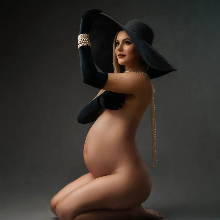 Kneeling expectant mother in wide black hat, velvet gloves and pearls; moody backdrop.