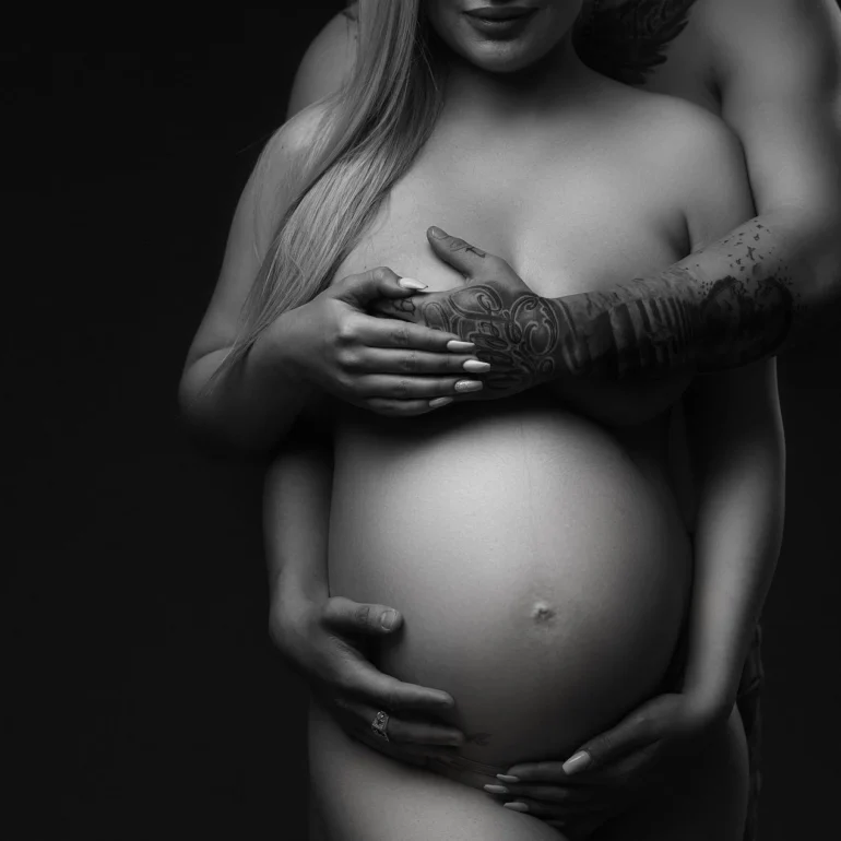 Close‑up B&W embrace; tattooed arms and hands over belly.