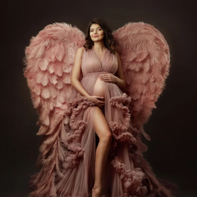 Mother‑to‑be in blush ruffled gown with pink angel wings; dreamy studio.